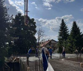 Construction workers pouring concrete on a building site with trees and a house in the background.